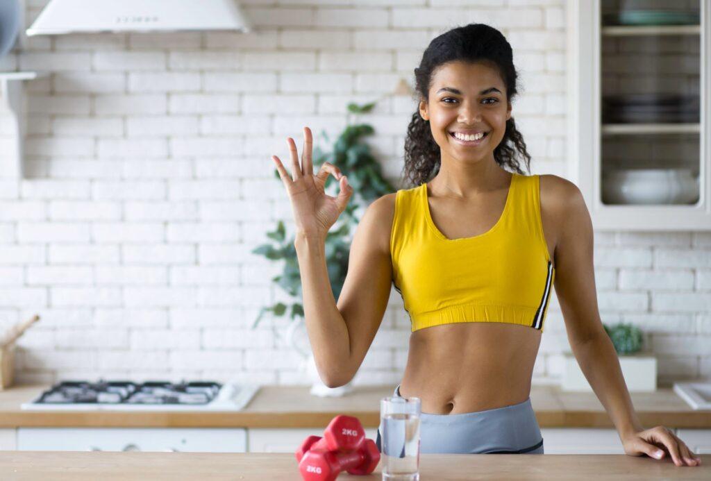 Fitnesstrainerin zeigt Geste OK, stehend in der Küche zu Hause, Blick in die Kamera, gesunder Lebensstil, Fitness zu Hause online